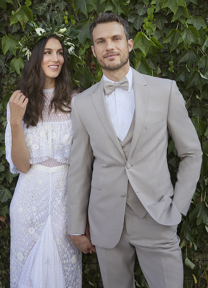 A Handsome man in a tan 'Aruba' Suit by Allure Men stands with a beautiful woman in a white dress in front of a wall of green ivy.