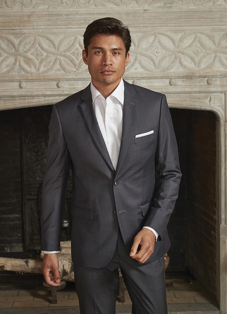 A man in a grey suit stands in front of a fireplace.