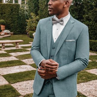 A handsome black man stands on a checkered garden clasping his hands while wearing a sea glass green 'brunswick' tuxedo.