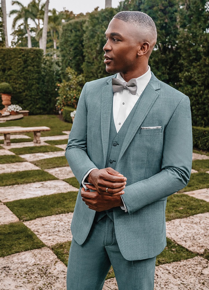 A handsome black man stands on a checkered garden clasping his hands while wearing a sea glass green 'brunswick' tuxedo.