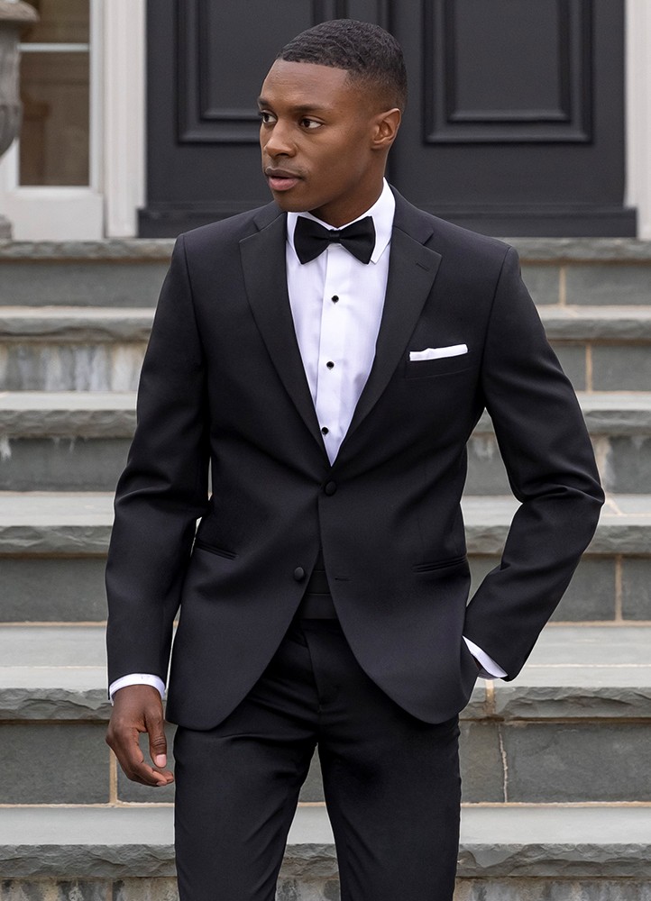 A handsome black man wearing the black 'Quincy' tuxedo walks down a flight of concrete stairs in front of a gorgeous mansion with his hand causally in one pocket.